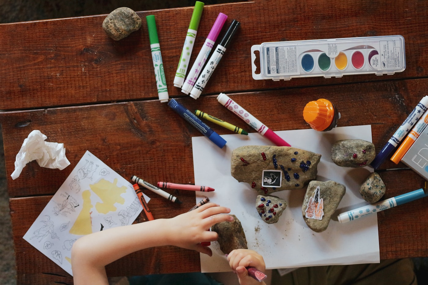 A child's hands decorating craft rocks with markers and paint at a wooden table