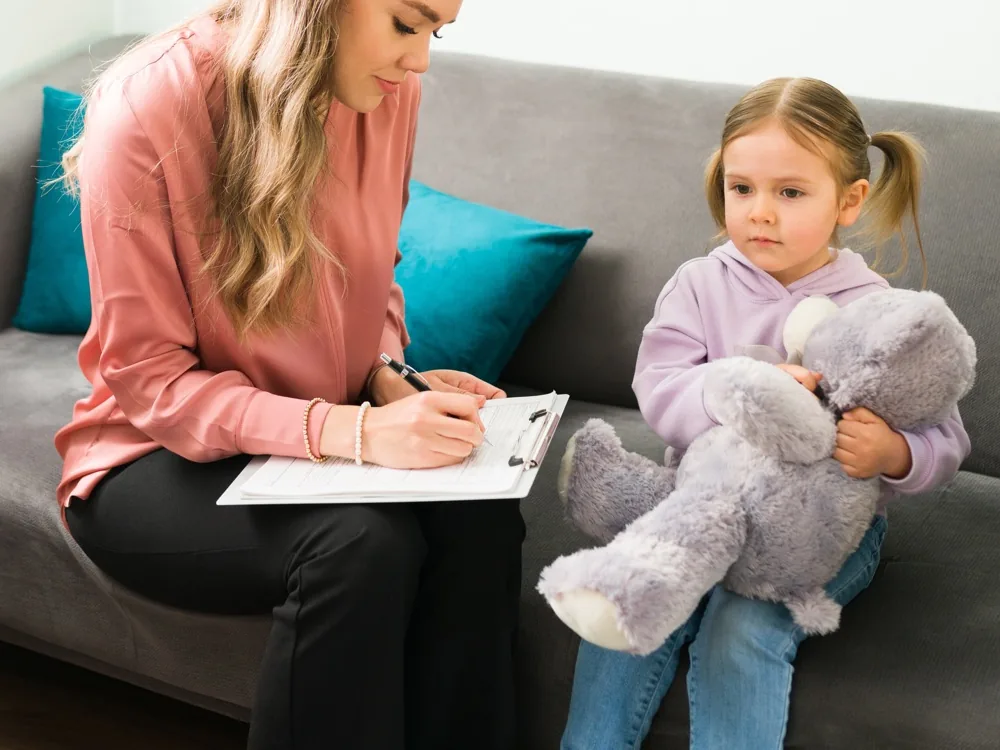 A clinician taking notes on a clipboard beside a young child holding a stuffed animal on a living-room sofa